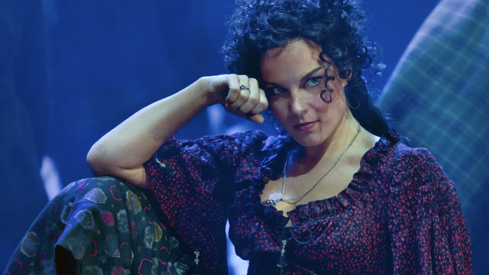 Actress in stage performance of Carmen sits with her elbow on a bent knee and her head resting on her fist
